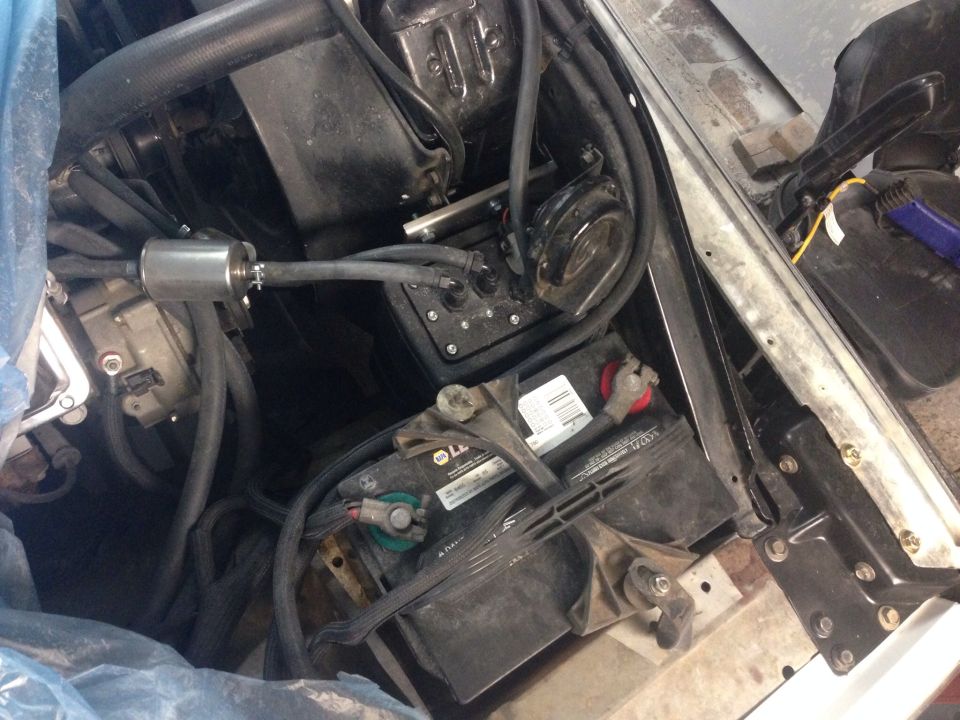 Car battery, fuel filter, and some hoses in an engine bay. Some blue plastic is covering parts