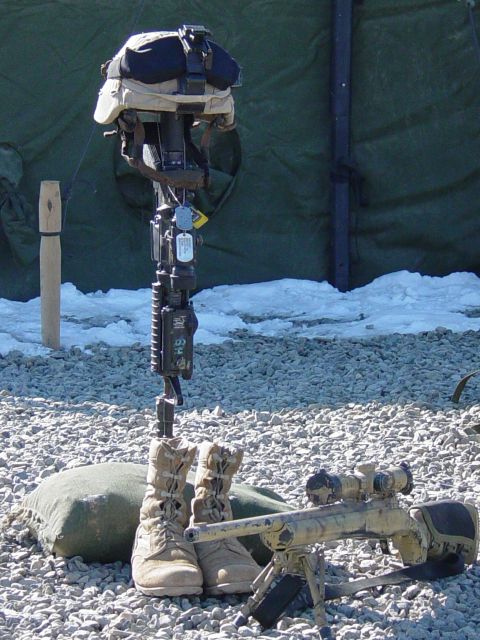 1981 Ford Bronco - A battlefield cross memorial, a helmet atop a rifle, accompanied by boots and a sniper rifle, stands in a gravelly, snowy outdoor setting.