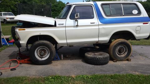 White and blue Ford Bronco on jack stands with hood open, another vehicle in background