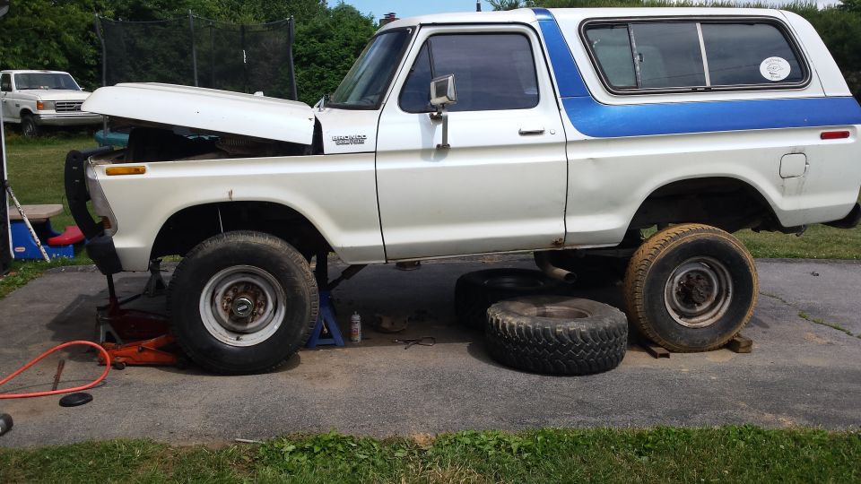 White and blue Ford Bronco on jack stands with hood open, another vehicle in background