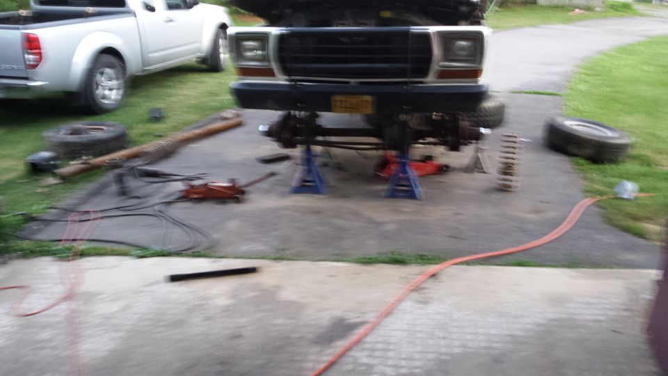 Ford truck on jack stands with front end disassembled. Another pickup truck is parked to the left