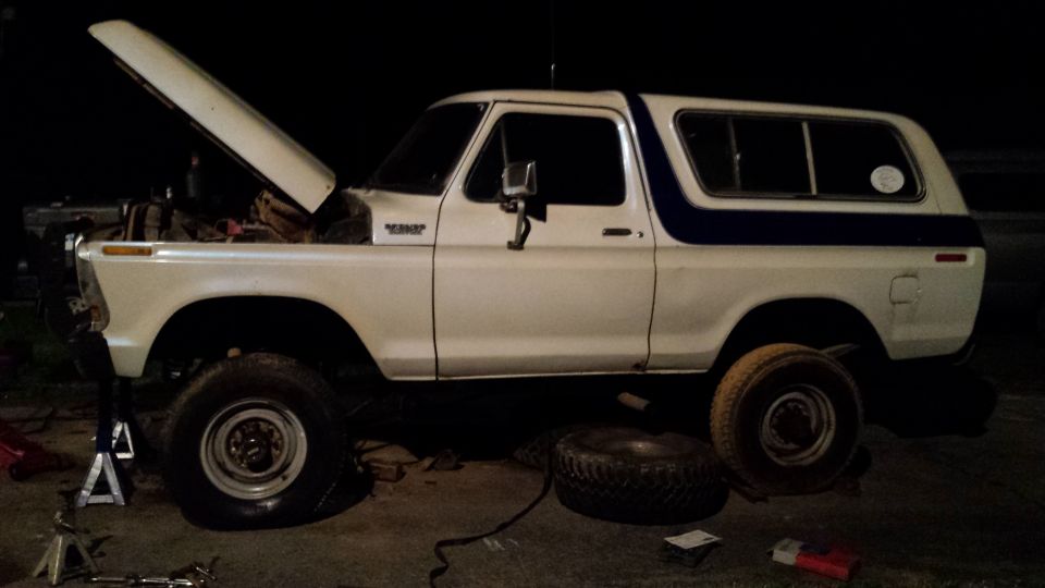 White 1978 Ford Bronco with blue stripe, hood open, on jack stands, with a spare tire on the ground