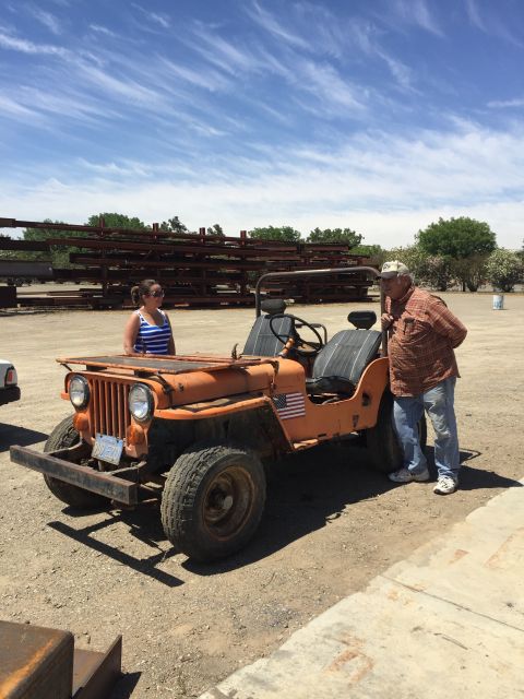 1946 Jeep Willys - An orange, open-top Jeep CJ5 is parked on a gravel lot under a blue sky with wispy clouds. A man and a woman stand next to the vehicle, whi…