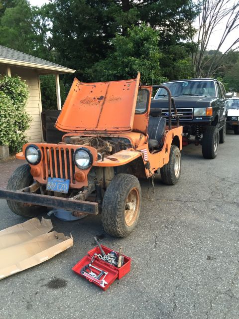 1946 Jeep Willys - An orange vintage Jeep CJ5 with its hood open is parked on an asphalt surface next to a black Ford Bronco. A red toolbox with various tools…