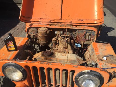 1946 Jeep Willys - An orange, rusted Jeep CJ5 is shown with its hood open, revealing the engine bay. A bottle of AutoZone Mercon V transmission fluid sits on…