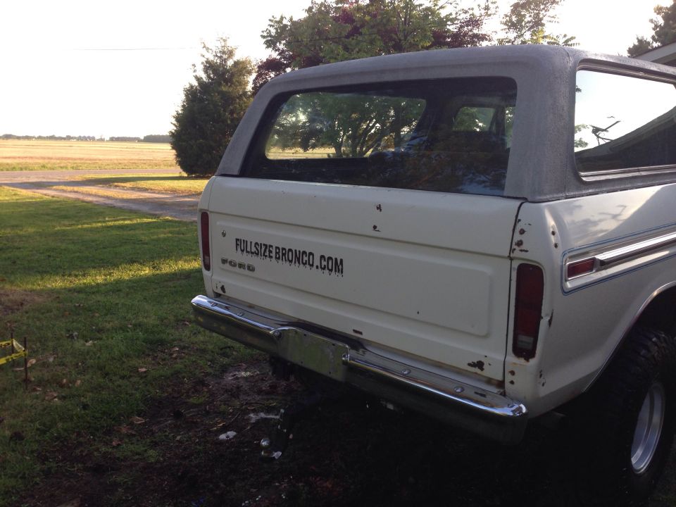 White Ford Bronco parked in a grassy field, showing the rear driver's side with a website decal.