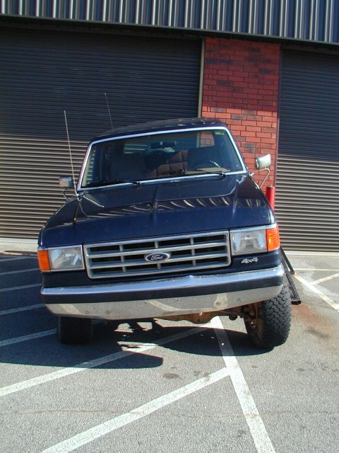 1988 Ford Bronco - A dark blue Ford Bronco is parked in front of a building with a brick wall and large garage doors.