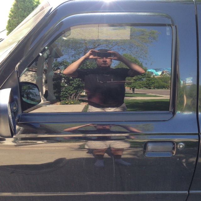 Reflection of a man taking a selfie in the window and door of a dark pickup truck.