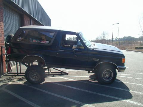 1988 Ford Bronco - A dark blue SUV, identified as a Bronco by its badge, is positioned on metal ramps in a parking lot.