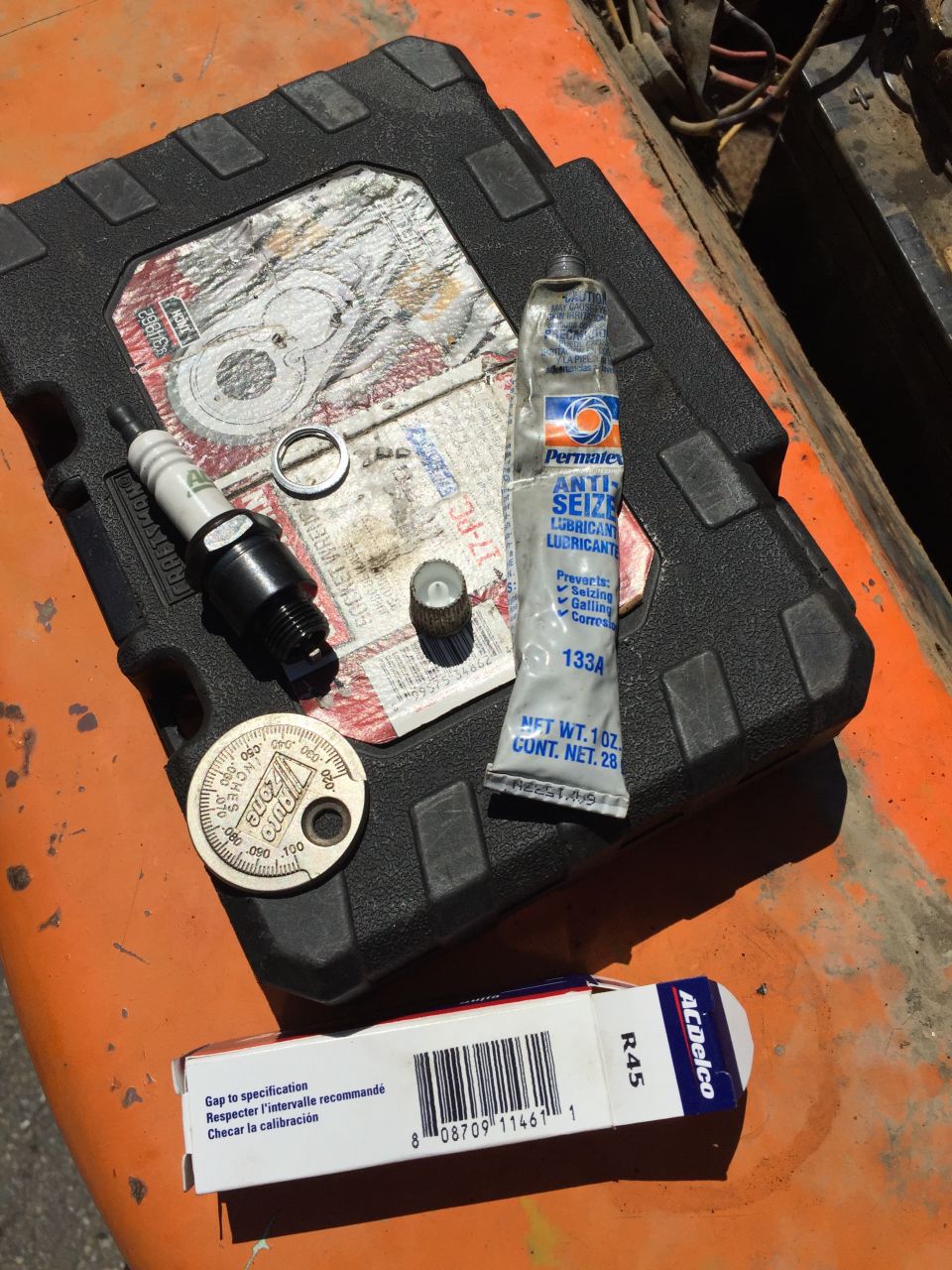 Spark plug, anti-seize, gap tool, and ACDelco spark plug box on orange surface.