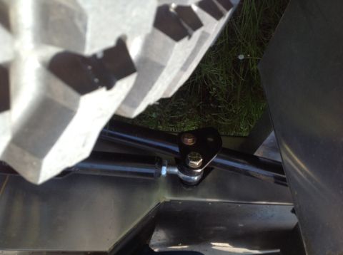 1992 Ford Bronco - A close-up view of a black metal tie rod assembly with two bolts attaching it to a bracket, mounted on a vehicle. A portion of a large, kno…