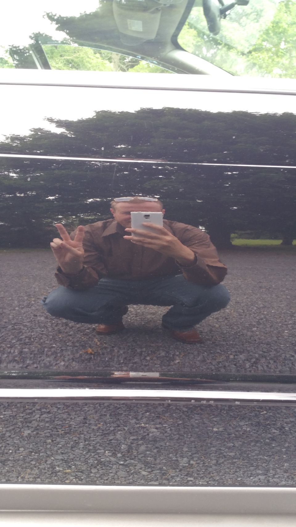 Man in brown shirt and blue jeans takes a selfie in the reflection of a dark car, crouching and making a peace sign.