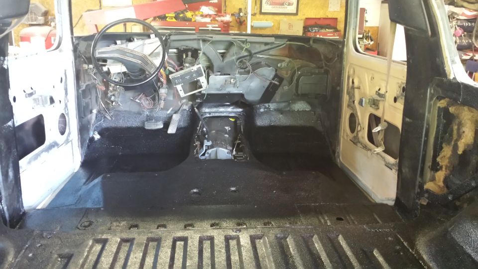 Interior of a classic Ford Bronco during restoration, showing dashboard, steering wheel, pedals, and black coated floor.