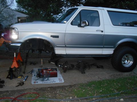 1994 Ford Bronco - A white and gray two-door Ford Bronco is parked outdoors with its front end elevated on jack stands. The front axle and suspension componen…