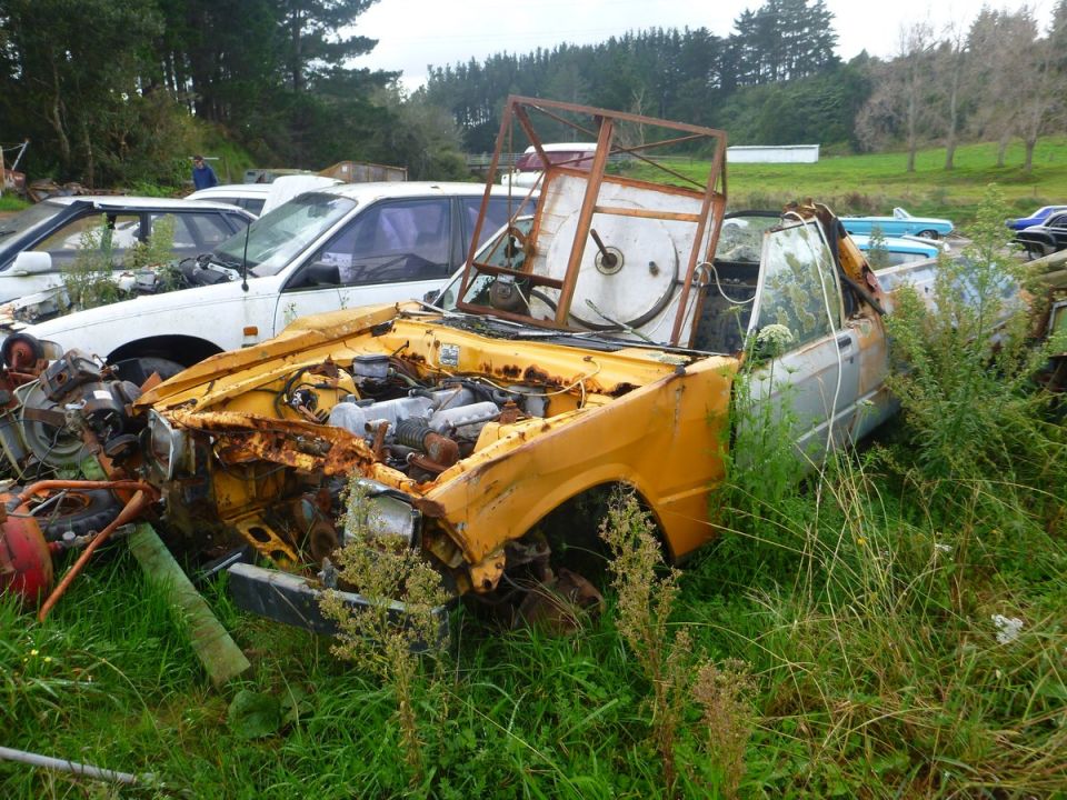 Rusted yellow and silver car with exposed engine in a grassy field.
