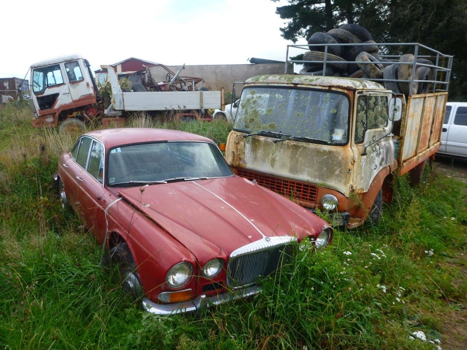 Red vintage sedan and rusty flatbed truck with tires in tall grass.