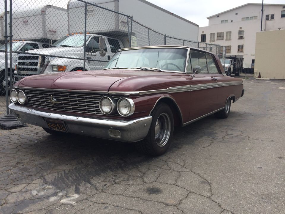 Maroon 1961 Ford Galaxie coupe parked on cracked asphalt, with white trailers and trucks behind a fence