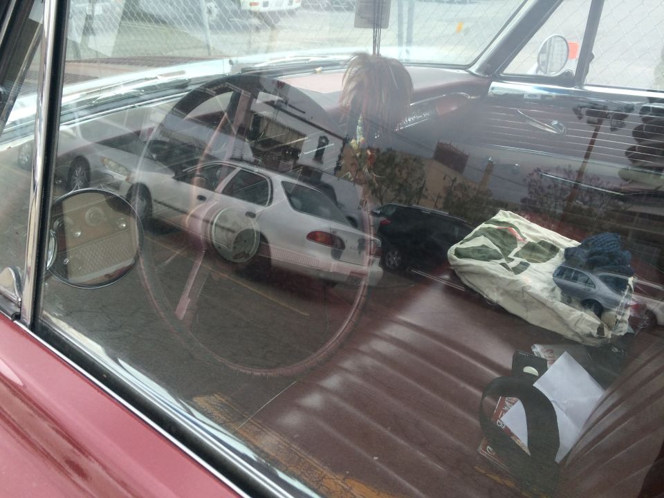 Interior of a red car with a large steering wheel and reflections of other cars and trees in the windshield