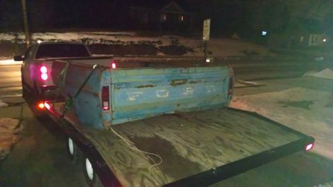 1994 Ford Bronco - A vintage Ford truck bed is transported on a trailer behind a newer pickup truck at night.