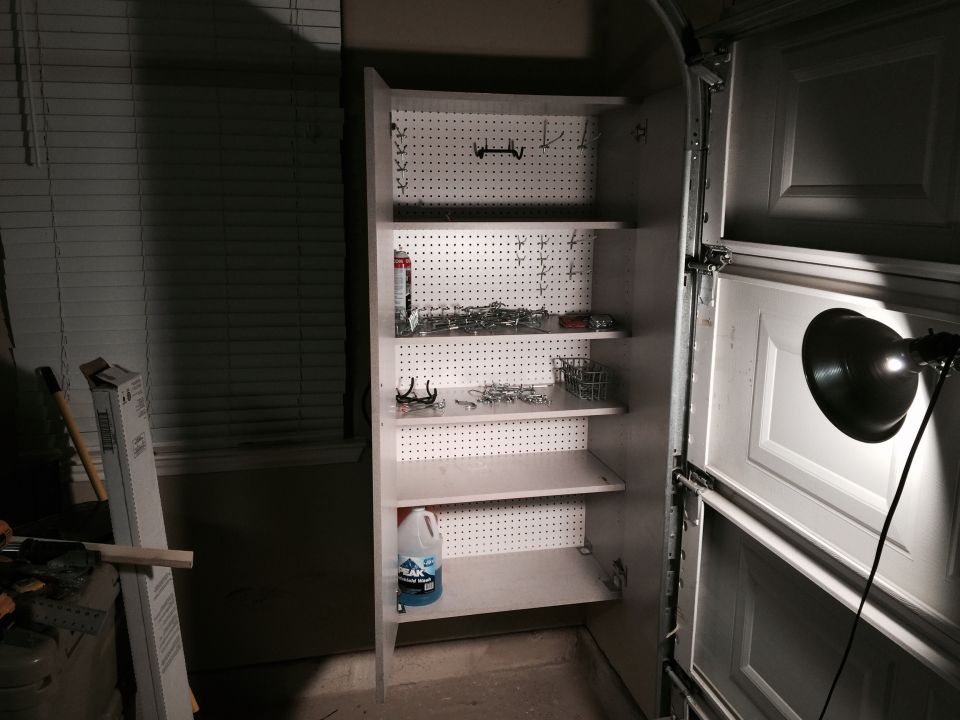 Garage storage cabinet with shelves holding various tools, hardware, and a bottle of cleaner. A garage door is partially open to the right