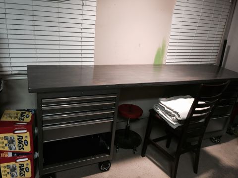 2015 Ford Deluxe - A workbench with a metal top and a rolling tool chest sits in front of a window with blinds. A chair with blankets is to the right.