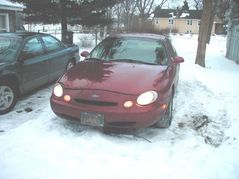 1997 Ford Taurus - A red Ford Taurus sits in the snow with its headlights illuminated, parked next to another vehicle.
