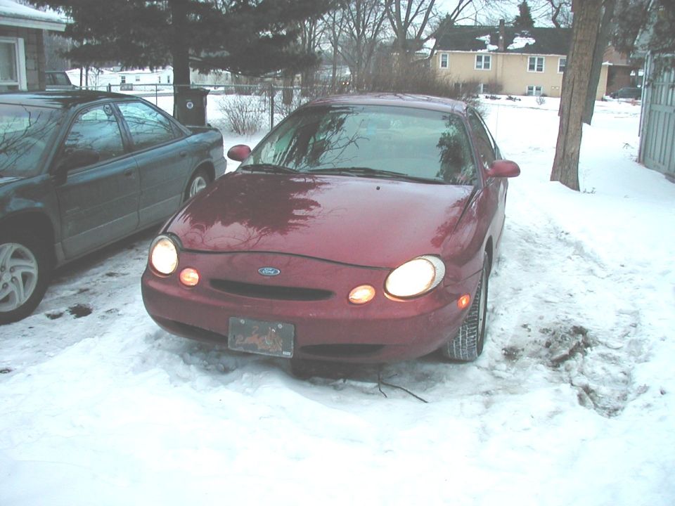 Red Ford Taurus parked in snow, front view. Headlights on, license plate visible