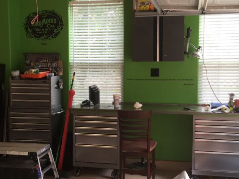 2015 Ford Deluxe - A cluttered garage workbench area features tool chests, supplies, and a vintage Ford Motor Co. logo on the green wall.