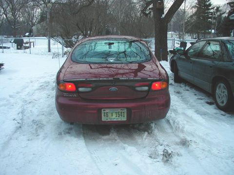 1997 Ford Taurus - The rear of a red 1997 Ford Taurus is visible in a snowy driveway next to another vehicle.