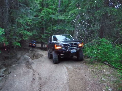 2005 Jeep Wrangler - Two dark-colored SUVs are driving on a dirt road through a dense forest. The front SUV has a light bar mounted on its bumper and a license…