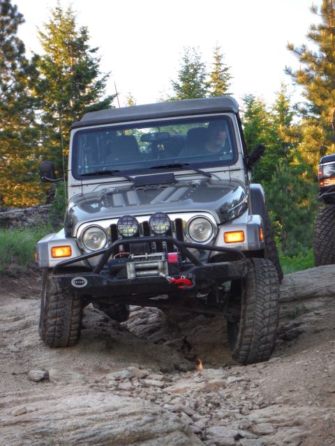 2005 Jeep Wrangler - A silver Jeep Wrangler is navigating a rocky, off-road trail with tall evergreen trees in the background. The front of the Jeep features a…