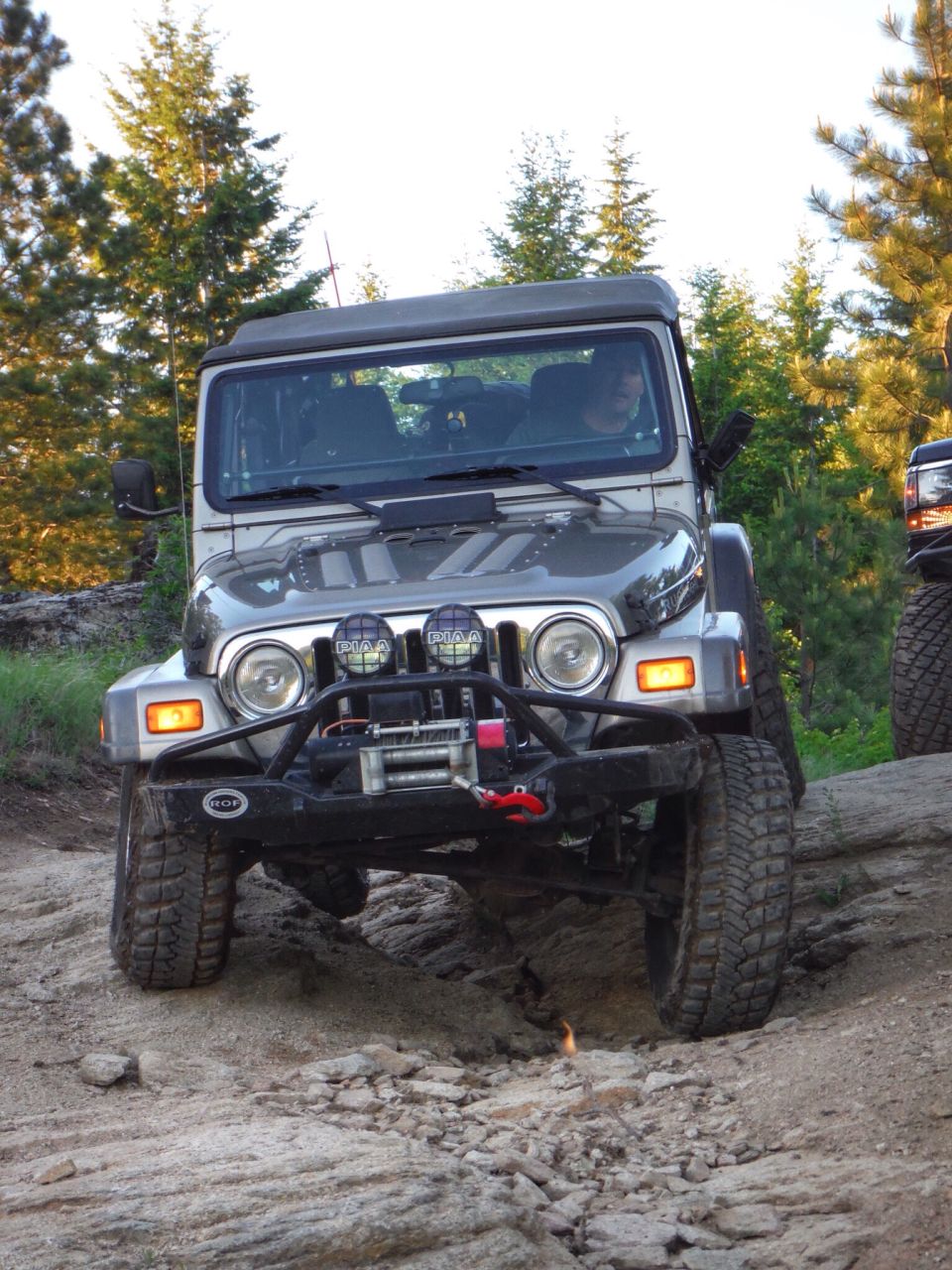 Silver Jeep Wrangler off-roading on a rocky trail with trees.