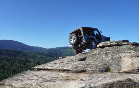 2005 Jeep Wrangler - A dark gray Jeep Wrangler is parked on a large rock formation with a forest and mountains in the background under a clear blue sky.