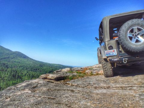 2005 Jeep Wrangler - A rear-view of a dark green Jeep Wrangler parked on a rocky overlook with a vast, tree-covered mountain range in the background under a cle…