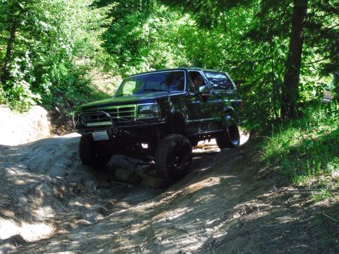 2005 Jeep Wrangler - A black SUV with a light bar on the front bumper is driving off-road on a dirt path surrounded by trees. The vehicle is angled on a slope w…