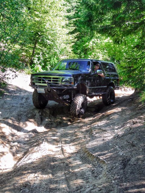 2005 Jeep Wrangler - A black Ford Bronco is driving off-road on a dirt path surrounded by trees. The vehicle has a lifted suspension and a custom front bumper.