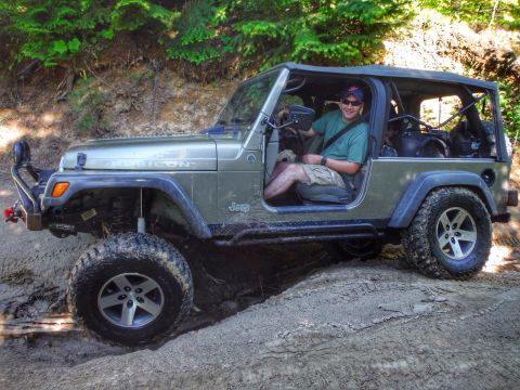 2005 Jeep Wrangler - A man wearing a green t-shirt and khaki shorts is driving a silver 2005 Jeep Wrangler Rubicon off-road on a dirt path.