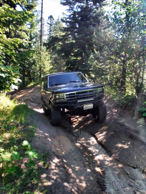 2005 Jeep Wrangler - A dark-colored SUV navigates a rocky, off-road trail through a sun-dappled forest. The vehicle has large tires, a light bar mounted on the…