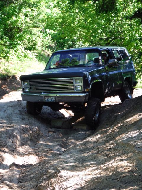 2005 Jeep Wrangler - A dark colored SUV is driving off-road on a sandy, rutted path surrounded by trees. The vehicle is angled towards the viewer as it navigate…