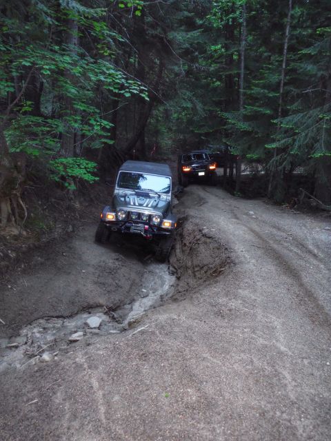 2005 Jeep Wrangler - A dark green Jeep Wrangler navigates a muddy, rutted trail in a dense forest, with a black SUV following behind.
