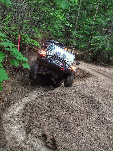 2005 Jeep Wrangler - A white Jeep Wrangler with off-road modifications navigates a muddy, rutted trail in a forest. Another dark-colored SUV is visible further…
