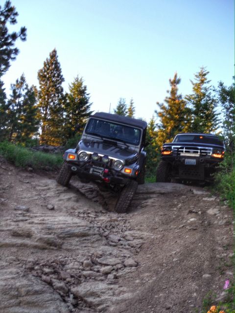 2005 Jeep Wrangler - Two off-road vehicles, a dark-colored Jeep Wrangler and a black pickup truck, navigate a rocky, uphill trail surrounded by evergreen trees.