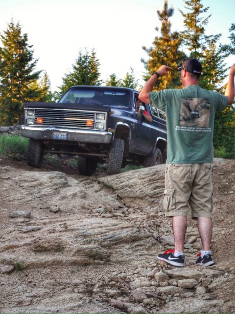 2005 Jeep Wrangler - A dark blue SUV is parked on a rocky incline with a man standing behind it, gesturing with his hands. The SUV has its headlights and turn s…