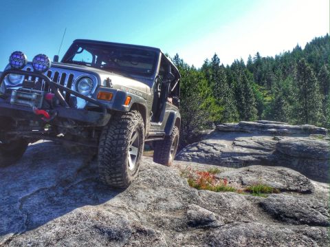 2005 Jeep Wrangler - A grey Jeep Wrangler Rubicon is parked on large rocks with a forest and clear blue sky in the background. The front of the Jeep is angled t…