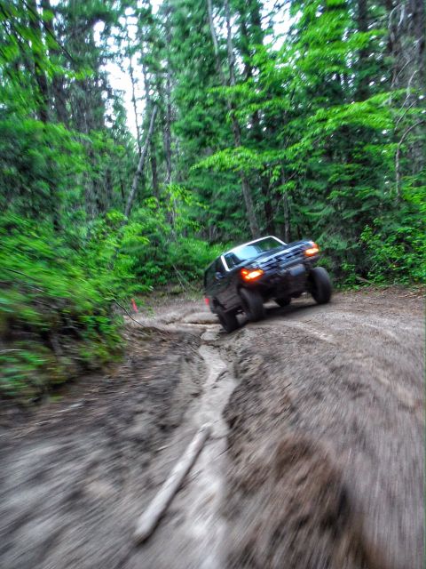 2005 Jeep Wrangler - A dark-colored Jeep Wrangler navigates a muddy, rutted trail through a dense forest. The vehicle is captured in motion, with motion blur in…