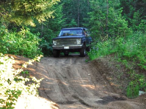 2005 Jeep Wrangler - A dark-colored, older model SUV drives up a dirt path in a wooded area. The vehicle is angled towards the viewer, with the front right side…