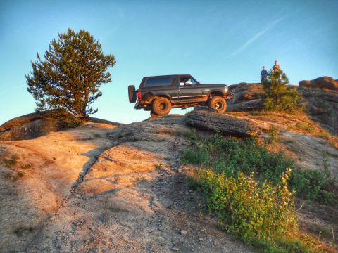 2005 Jeep Wrangler - A dark-colored SUV is parked on a rocky, elevated terrain with two people standing on the rocks behind it. A tree is visible to the left of…
