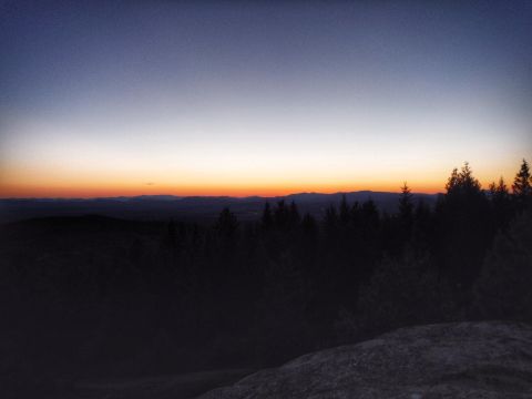 2005 Jeep Wrangler - The sky transitions from deep blue to orange and yellow hues as the sun sets behind a silhouetted mountain range and a dense forest.