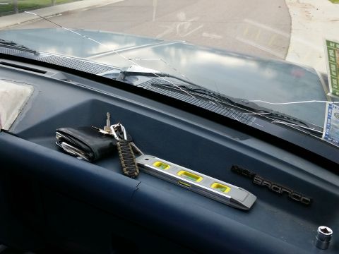 1988 Ford Bronco - A close-up view of a cracked windshield and dashboard of a vehicle. Items on the dashboard include a wallet, keys, a small level, and a Bro…