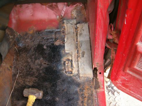1979 Ford Bronco - This image shows a heavily rusted interior floor pan of a red vehicle, with visible metal patches and welds indicating repairs.
